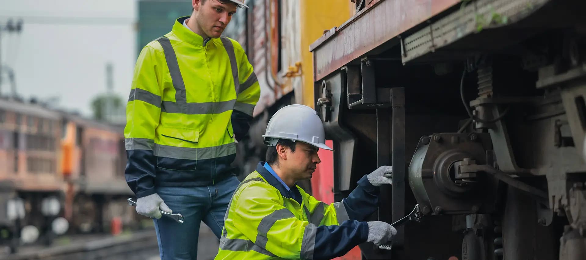 Two workers performing maintenance on a traincar. One worker is using a handheld tool to tighten a bolt, while the other is observing the process. The image highlights the need for efficient, reliable, and safe maintenance solutions in the rail industry.